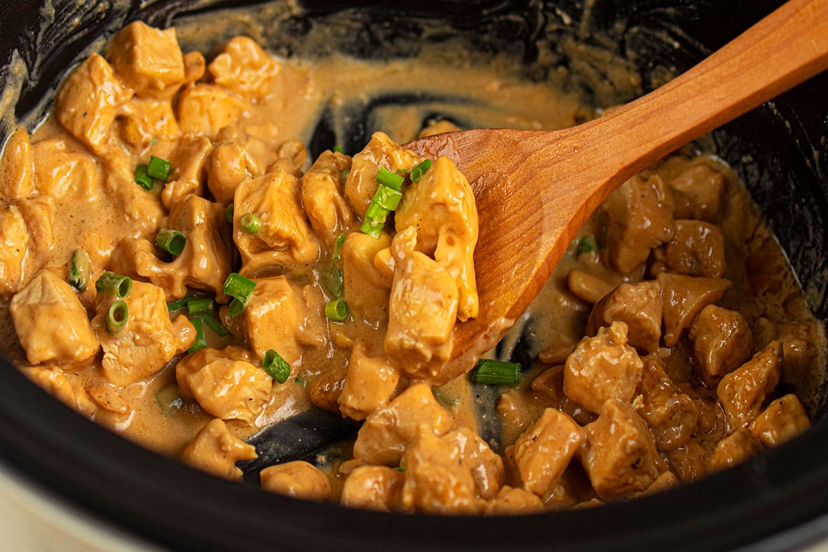 Black crockpot with nut based Asian chicken and creamy gravy along with large wooden spoon.