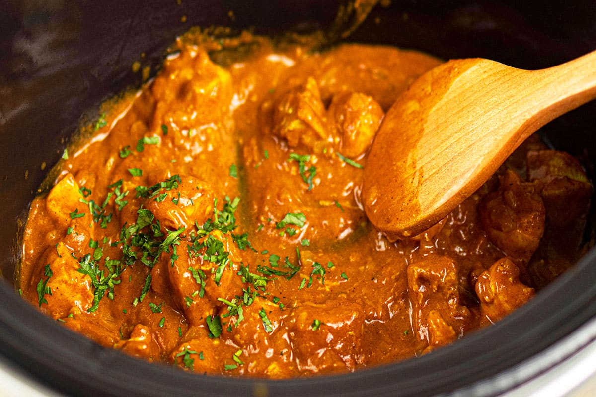 Easy homemade chicken curry in black slow cooker with wooden spoon.